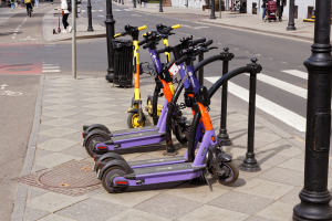 Eine Reihe von E-Scootern, die entlang einer Straße mit Fußgängern, Radfahrern, einem Müllcontainer im Vordergrund und Gebäuden, Bäumen und Pflanzen im Hintergrund geparkt sind.