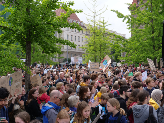 Eine große Menge von Menschen protestiert vor einem Gebäude in Berlin, hölt Schilder und steht in der Nähe von Bäumen, Fahrzeugen und einem Lautsprecher.
