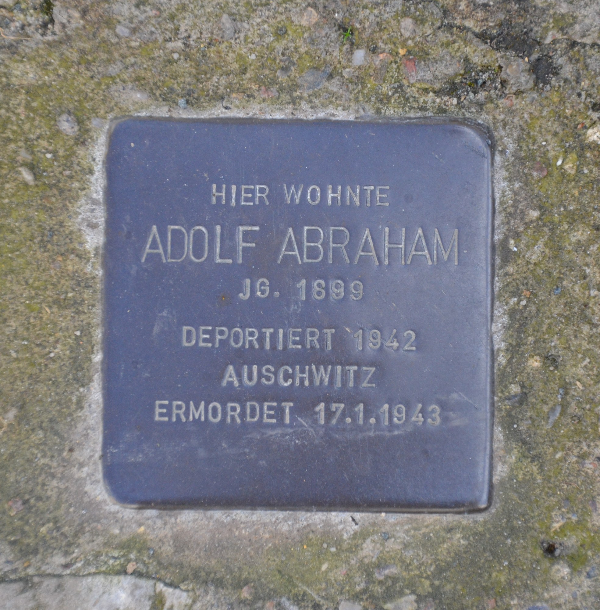 Rechteckige Plakette mit 'Adolf Abraham' eingraviert, an einer Steinwand angebracht.