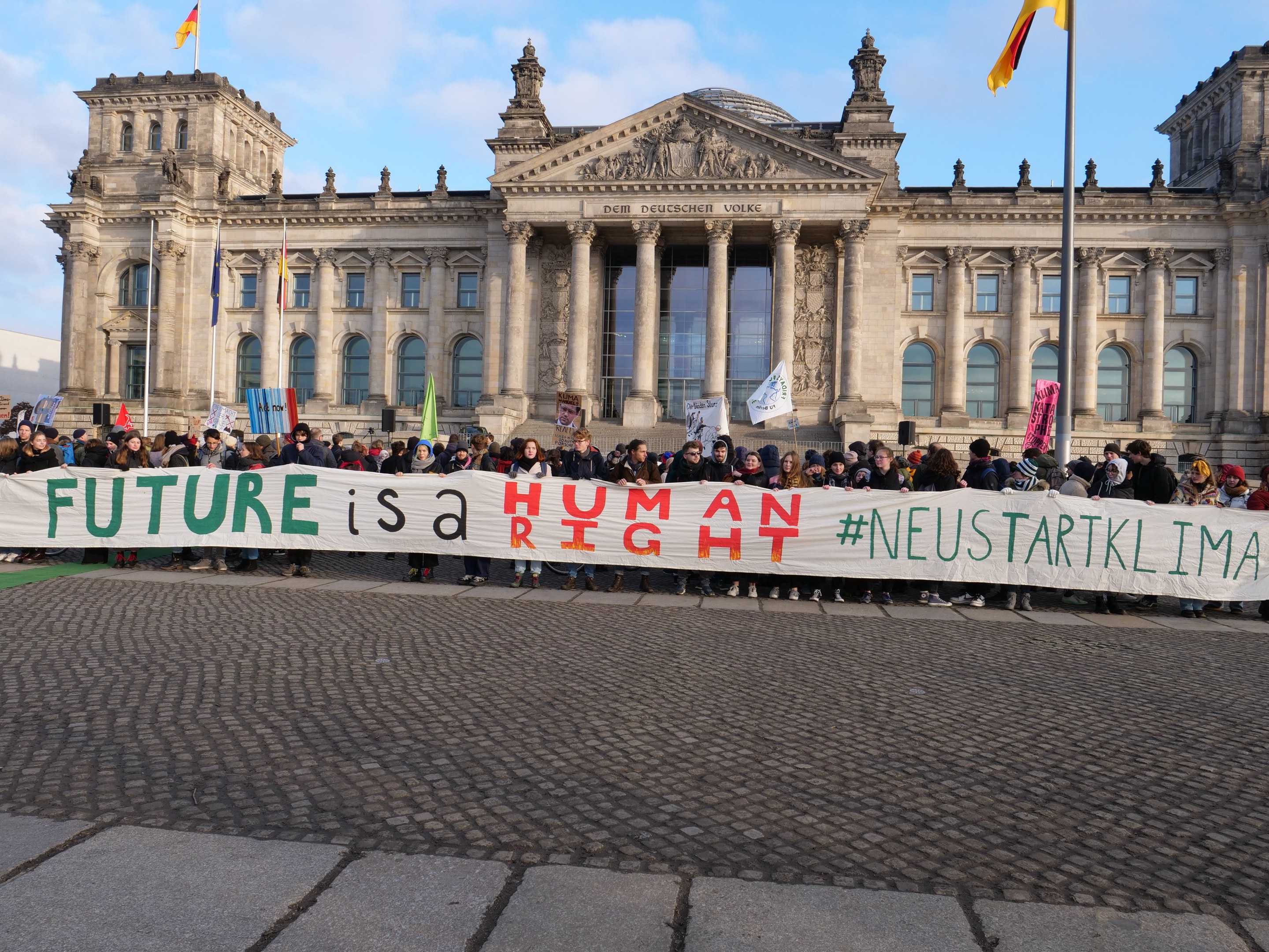 Gruppe von Menschen mit einem Banner mit der Aufschrift "Zukunft ist ein Mensch" vor dem Reichstagsgebäude in Berlin, Deutschland, mit bewölktem Himmel im Hintergrund.
