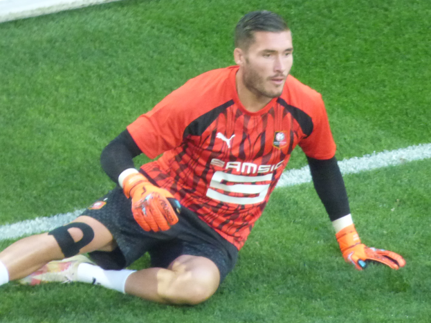 Ein Fußballspieler in rotem T-Shirt, schwarzen Shorts, weißen Socken und orangen Baseballhandschuhen sitzt auf dem Boden mit den Händen auf den Knien.