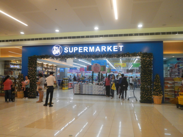 Eine Gruppe von Menschen vor einem Supermarkt, einige halten Einkaufstaschen, mit einem Einkaufswagen, einem Topf mit einer Pflanze, Regalen, Schildern und Deckenleuchten im Hintergrund.