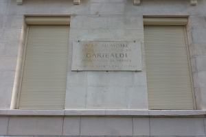 Ein Gebäude mit zwei Fenstern und geschlossenen Fensterläden sowie einer Gedenktafel an der Seite.