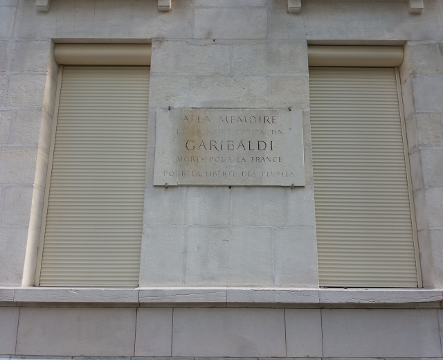 Ein Gebäude mit zwei Fenstern und geschlossenen Fensterläden sowie einer Gedenktafel an der Seite.