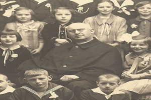 Ein altes Schwarz-Wei├č-Foto eines strengen Mannes, der in der Mitte sitzt und von einer Gruppe Kinder umgeben ist, die ihn bewundernd anschauen.