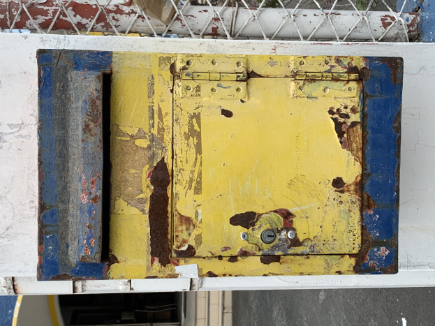 Ein gelb-blauer Metalltresor steht an einer weißen Wand mit einem Metallzaun im Hintergrund.