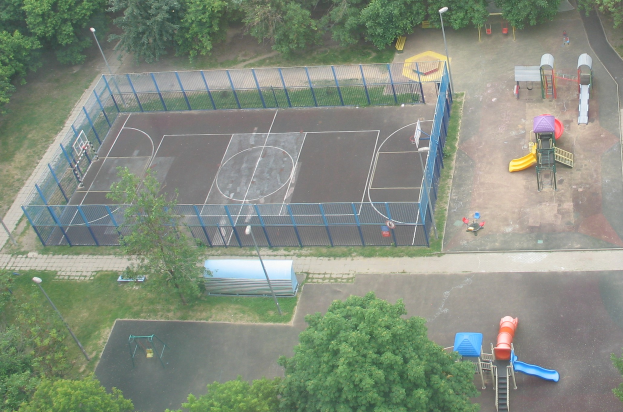 Eine Luftaufnahme eines Parks mit einem eingezäunten Basketballplatz, Straßenlaternen, Bäumen, Gras, Rutschen und anderem Spielplatzgerät.