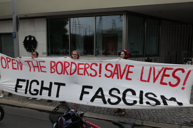 Demonstranten halten ein Transparent mit der Aufschrift "Öffne die Grenzen, rette Leben, bekämpfe Faschismus" vor einem Gebäude mit Glasfenstern und einem Mast, mit einem Fahrrad und einer Tasche auf der Straße.