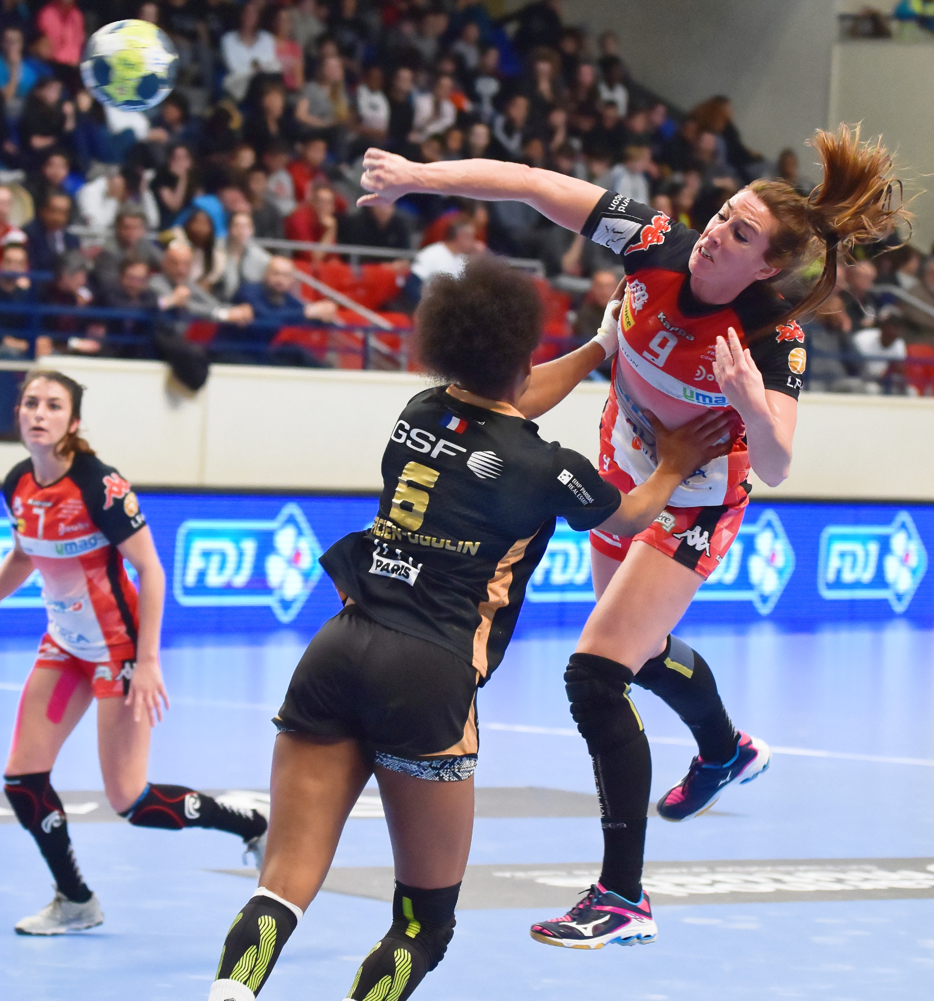 Eine Gruppe von Frauen spielt Handball auf einem Feld mit einem Ball in der Luft, während Zuschauer von Stühlen, Geländern und einer Wand im Hintergrund zuschauen.