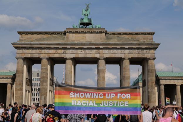 Eine Gruppe von Menschen hält eine "Racial Justice"-Schleife vor dem Brandenburger Tor in Berlin, Deutschland, mit Gebäuden und einem bewölkten Himmel im Hintergrund.