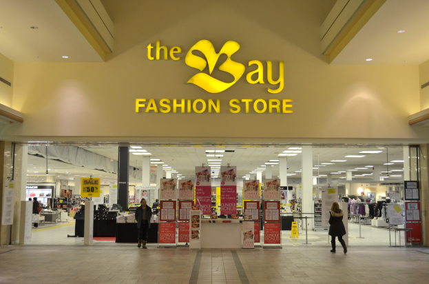 Außenansicht eines Bay Fashion Store in einem Einkaufszentrum mit Einkäufern, Wandtexttafeln, Deckenleuchten, Säulen und Hintergrundauslagen.