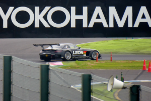 Schwarzer Sportwagen auf einer Rennstrecke mit einem "Yokohama"-Schild im Hintergrund, umgeben von Verkehrskegeln, Gras und einem Zaun, mit einem Lautsprecher im Vordergrund.