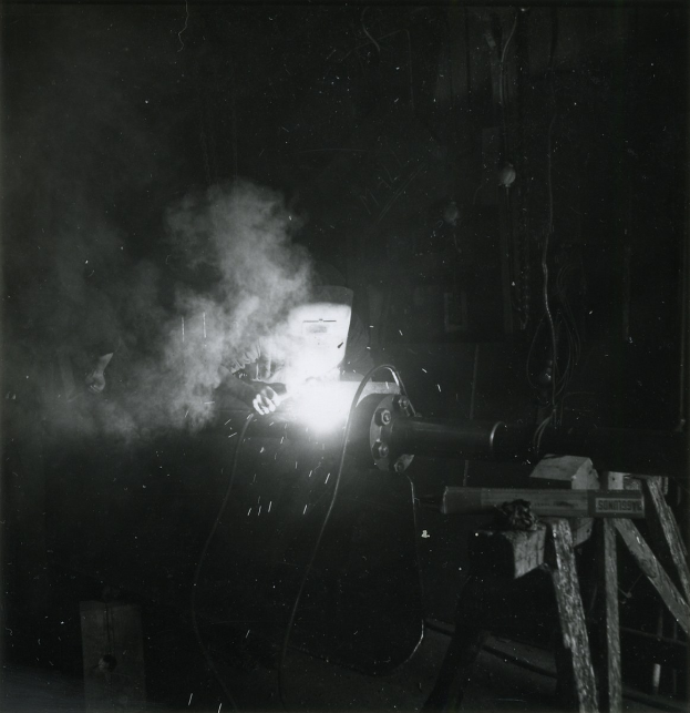 Schwarzes und weißes Foto eines Schweißers, der an einem Metallstück arbeitet, mit Rauch, der aus der Schweißmaschine aufsteigt und verschiedenen Gegenständen im Hintergrund.