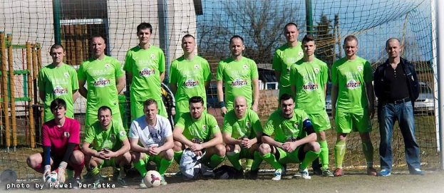 Eine Gruppe von Männern in grünen T-Shirts steht auf einem Fußballfeld mit einem Ball davor, einige sind in der Hocke, in der Nähe eines Torpfahls mit Bäumen, Gebäuden und Fahrzeugen im Hintergrund.