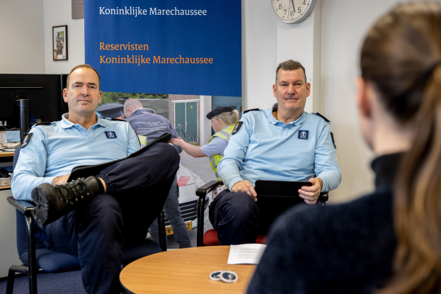 Zwei Polizeibeamte sitzen an einem Tisch und sprechen mit einer Frau; im Hintergrund sind eine Wandtafel, eine Uhr und Fotorahmen zu sehen, sowie ein weiterer Tisch mit einem Monitor, Tastatur und Maus links daneben.