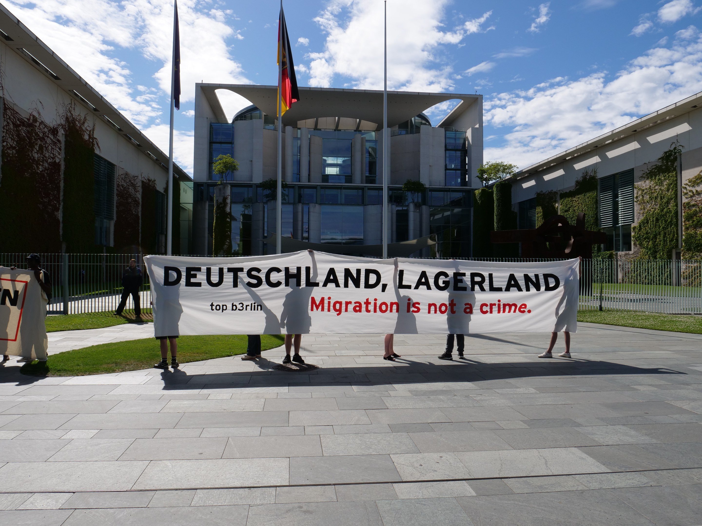 Eine Gruppe von Menschen mit einer Fahne mit der Aufschrift "Deutschland, Lagerland Migration ist kein Verbrechen" vor einem Gebäude mit Fenstern, umgeben von Gras, einem Metallzaun, Pflanzen, Bäumen und Fahnenstangen unter einem bewölkten Himmel.