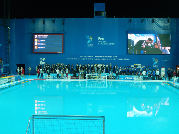 Gruppe von Menschen, die um ein Hallenbad mit Netz am Boden stehen, mit einer Wand im Hintergrund, die "FINA World Championships 2015 Kazan" auf einem Bildschirm anzeigt, sowie Trägern und Lampen im Hintergrund.