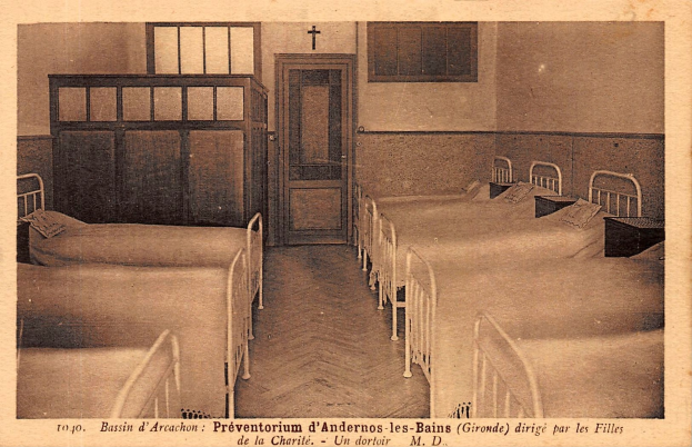 Ein altes Foto eines Krankenhauszimmers mit Betten, Kissen, Tischen, einer Tür, Fenstern und einem Kreuz an der Wand, mit Text am unteren Bildrand.