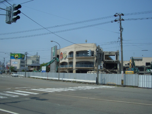 Eine Stadtstraße mit Fahrzeugen, Strommasten, Laternen, einer Ampel auf der linken Seite, Gebäuden, Bäumen und dem Himmel im Hintergrund und einem abbruchreifen Gebäude im Vordergrund.
