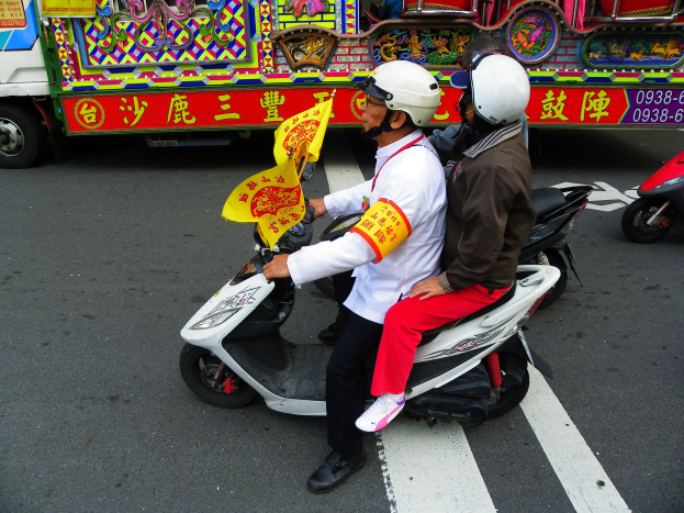 Zwei Personen mit Helmen fahren auf einem Motorroller, eine hölt eine gelbe Flagge, mit einem Fahrzeug im Hintergrund.