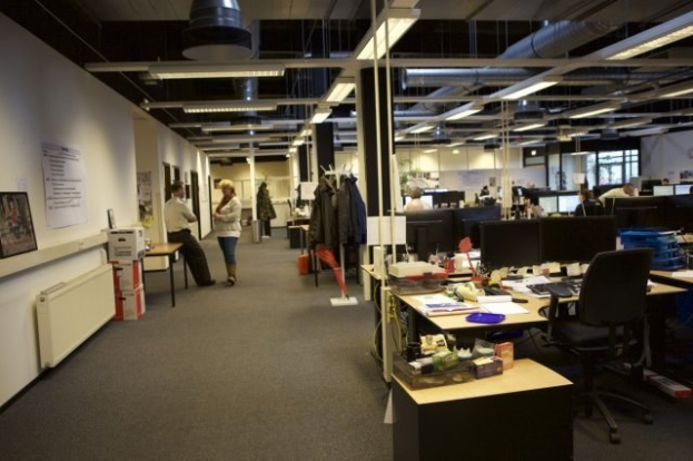 Gro├če offene Bürolandschaft mit Menschen, die an Tischen mit Computern, Monitoren, Tastaturen und Papieren arbeiten, umgeben von Stühlen, Kisten und Büchern; Klamotten h├Ąngen von Gestellen, W├Ąnde zeigen Tafeln mit Text und Bildern und Deckenlampen erhellen den Raum mit Glasfenstern im Hintergrund.