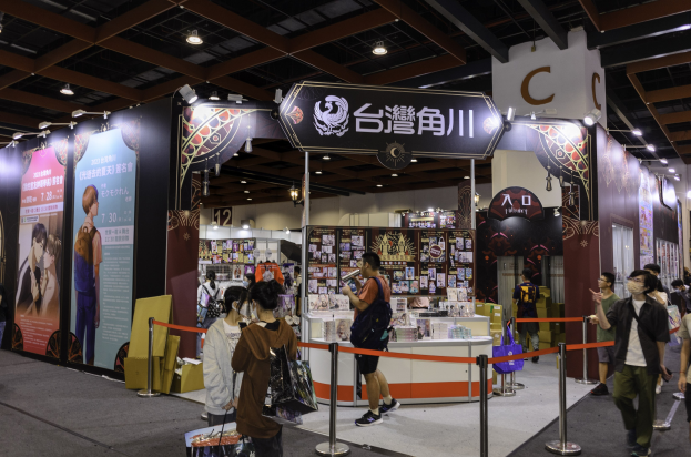 Eine Gruppe von Menschen steht vor einem Messestand, einige halten Taschen, mit Bändern, Informationsplakaten, Deckenleuchten und Ausstellungsregalen im Hintergrund, wahrscheinlich für die Tokyo Anime Expo 2019.