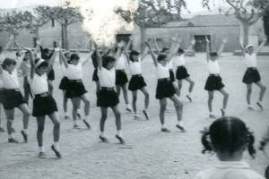 Schwarzes und weißes Foto von jungen Mädchen in Schuluniformen, die auf einer Straße tanzen, die von Gebäuden und Bäumen gesäumt ist, mit einem Paar Schuhe am unteren Rand.