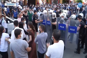 Eine Gruppe von Menschen steht vor einer Reihe von Polizisten mit Helmen und Schilden, mit einem Auto links, einer Person mit einer Kamera rechts und Geb├Ąuden, B├Ąumen und Texttafeln im Hintergrund in Ankara, T├╝rkei.