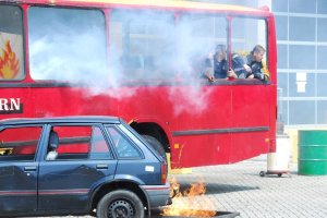 Roter Doppeldeckerbus mit Rauch mit drei sichtbaren Passagieren, neben einem Auto geparkt, vor einem Glasfenster-Gebäude mit einem Fass auf der rechten Seite.