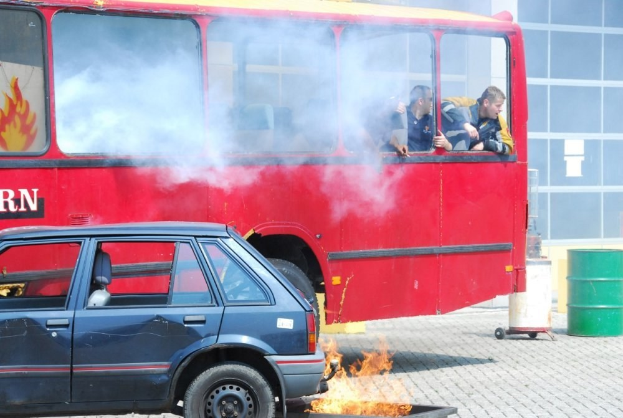 Roter Doppeldeckerbus mit Rauch mit drei sichtbaren Passagieren, neben einem Auto geparkt, vor einem Glasfenster-Gebäude mit einem Fass auf der rechten Seite.