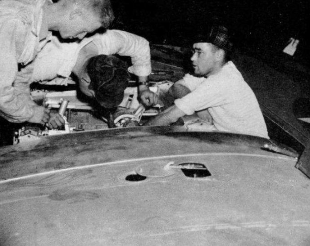 Drei Männer in Mützen arbeiten an einem schwarz-weißen Auto in einer Garage, umgeben von Werkzeugen und Materialien.