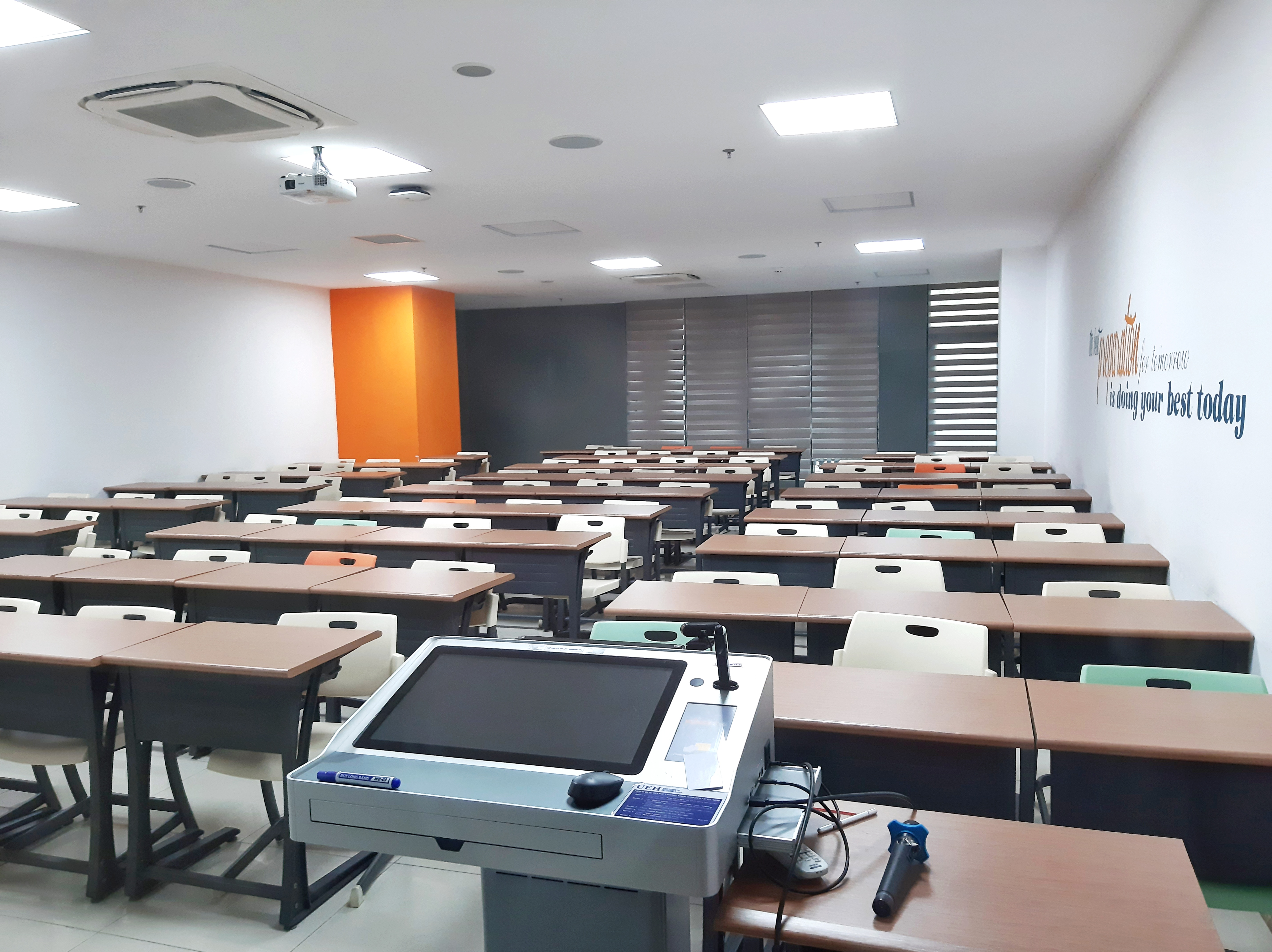 Ein Klassenzimmer mit Tischen und Stühlen in Reihen, einen Deckenprojektor und verschiedene Gegenstände auf den Tischen sowie Text an der rechten Wand, der darauf hinweist, dass es sich um eine Privatschule in Singapur handelt.