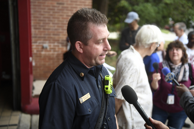 Polizeibeamter spricht zu einer Gruppe von Menschen mit Mikrofonen, Bäumen und einem Gebäude im Hintergrund.