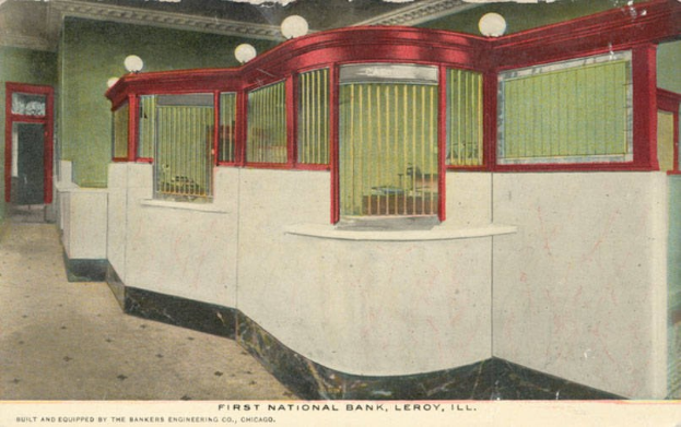 Schwarz-weiß-Postkarte vom Inneren der ersten Nationalbank in Leroy, Illinois, die einen Raum mit einer Wand, Fenstern, Lampen, einer Tür und einem Boden zeigt, mit Text unten.