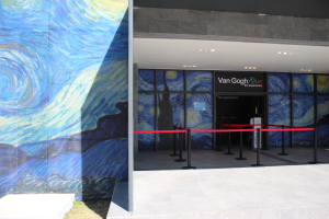 Der Eingang eines Gebäudes zeigt ein großes Wandgemälde, ein Schild mit der Aufschrift "Van Gogh Alive", Absperrpfosten mit Bändern, Gras auf dem Boden und Lampen an der Decke.