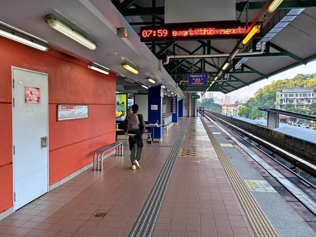 Ein Mann geht über einen Bahnsteig und trägt eine Tasche, in der Nähe eines Zuges mit Bänken, Leuchten, Informationsanzeigen und einer Tür, mit Bäumen, Gebäuden und Himmel im Hintergrund.