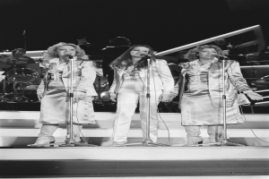 Drei Frauen singen in Mikrofone auf der Bühne mit Musikern im Hintergrund, schwarz-weiß.