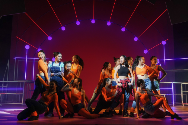 Eine Gruppe junger Tänzer in bunten Kostümen performs auf einer Bühne mit bunten Lichtern und Geländern, mit erhobenen Armen, während das Publikum jubelt und klatscht.