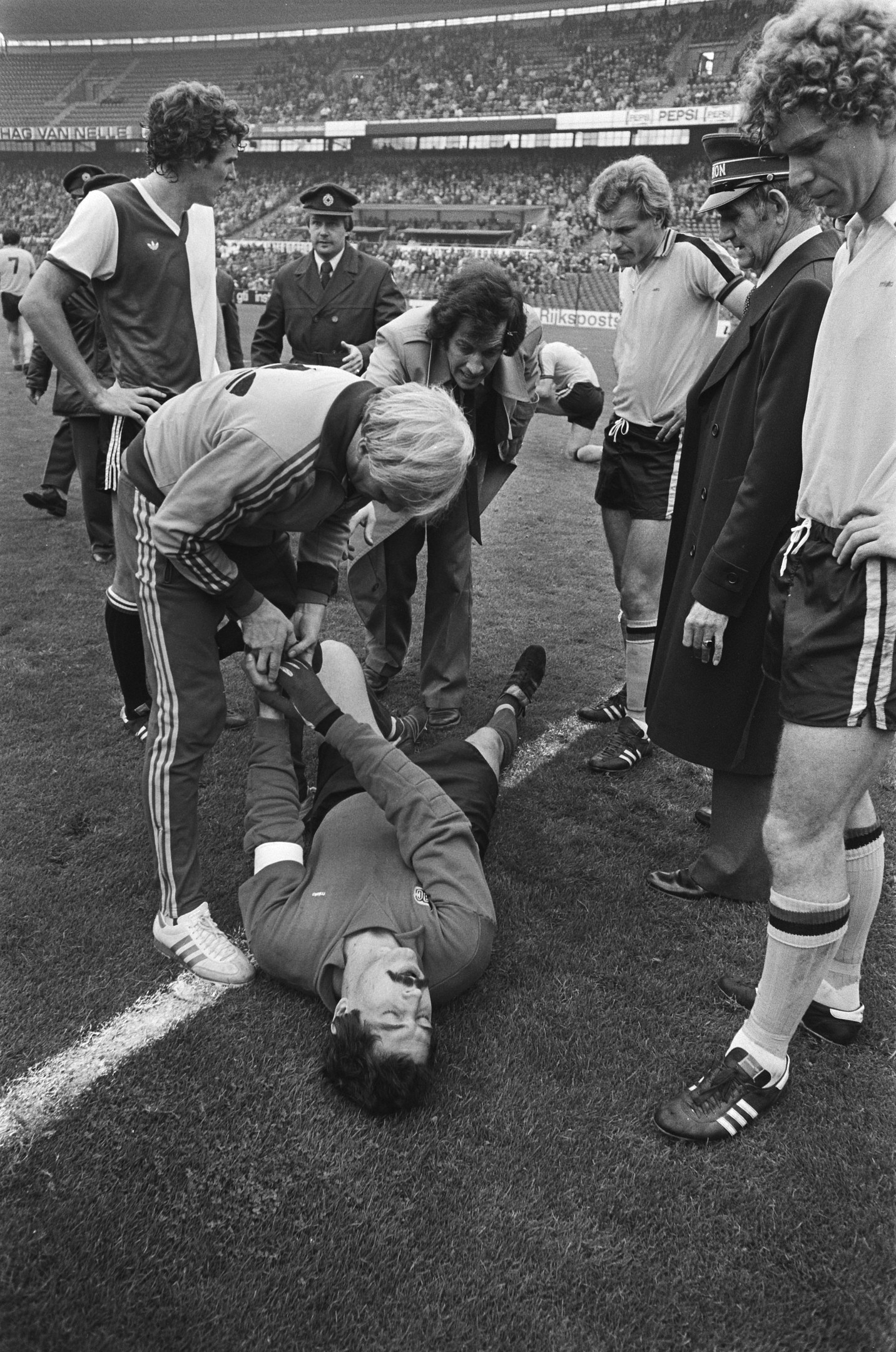 Schwarzes und weißes Bild eines Fußballspielers, der auf dem Boden liegt und von Mannschaftskameraden und einem Stadionpublikum umgeben ist und verletzt zu sein scheint.