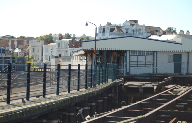 Ein Bahnhof mit einem Zug auf den Schienen, eine Brücke mit Geländern, Laternen, Verkehrsampeln mit Masten, Gebäude mit Fenstern, Bäume und ein Himmel im Hintergrund.