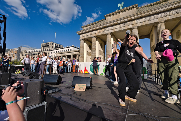 Eine Gruppe von Menschen auf einer Bühne mit Mikrofonen und Kameras vor dem Brandenburger Tor, mit Lautsprechern und Equipment im Hintergrund, vor Gebäuden, Bäumen und Himmel.