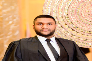 Ein Mann in schwarzer Robe, weißem Hemd und schwarzer Krawatte steht vor einer goldenen und cremefarbenen Wand, dargestellt als "Richter des Jahres".