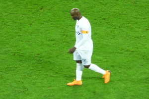 Ein Fußballspieler in einem weißen T-Shirt, weißen Shorts und orangefarbenen Schuhen läuft über ein saftig grünes Feld.