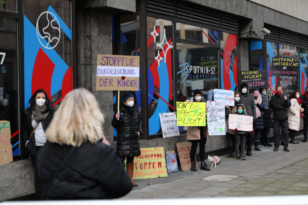 Maskentragende Menschen mit Schildern und Plakaten vor einem Gebäude mit Glasfront, mit Kameras und einem Hund, der scheinbar gegen das Verbot von Schulmasken durch die deutsche Regierung protestiert.