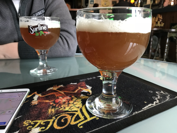 Zwei Gläser goldenes Bier auf einem Tisch neben einem Handy, mit Menschen und gerahmten Wandbildern im Hintergrund.