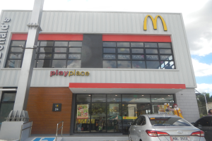 Ein McDonald's-Restaurant mit parkenden Autos davor, umgeben von Bäumen und Pfählen unter einem klaren blauen Himmel, mit Glaswänden, Fenstern und einem "McDonald's Playplace"-Schild, mit sichtbaren Postern und Gegenständen im Inneren.