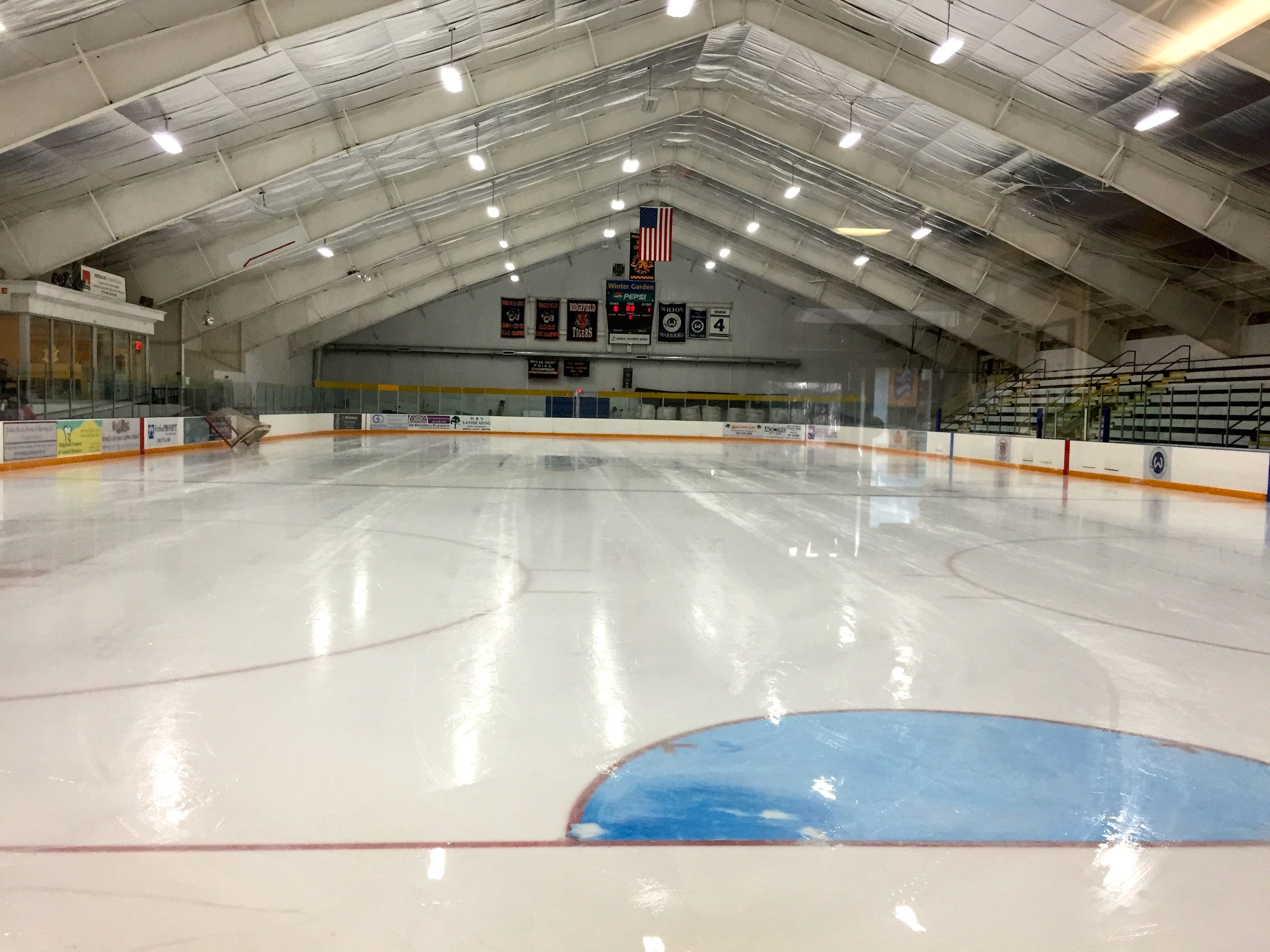 Großes Indoor-Eisstadion mit Deckenbeleuchtung, Werbetafeln, Treppen mit Geländern, einer Wand im Hintergrund und einigen Menschen, was darauf hinweist, dass es sich um eine Sportanlage handelt, die likely für Hockey verwendet wird.