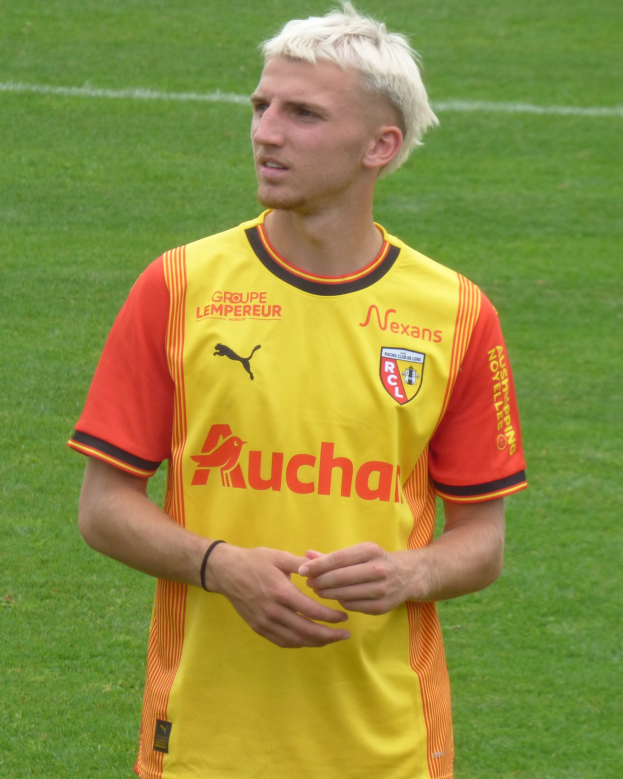 Ein Fußballspieler in einem gelb-orangen gestreiften Trikot steht selbstbewusst auf einem Feld und scheint ein Flügelspieler mit entschlossener Miene zu sein.