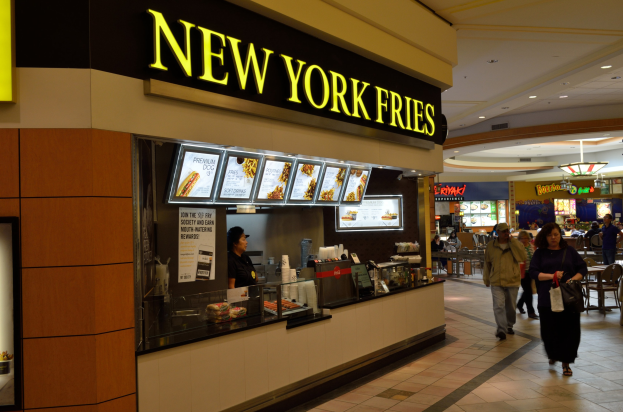 Ein New York Fries Restaurant in einem Einkaufszentrum mit Menschen, die an Tischen sitzen und gehen, eine Theke mit verschiedenen Gegenständen, eine Tafel mit Text auf der linken Seite und Tafeln mit Text und Food-Images im Hintergrund, alles unter einer Decke mit Lampen.