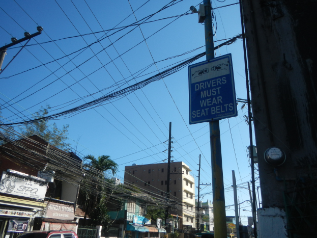 Stadtstraße mit fahrenden Autos, Strommasten mit Drähten, Gebäude, Bäume und Namensschilder, mit einem "Fahrer müssen Sicherheitsgürtel tragen"-Schild an einem Strommasten vor einem sichtbaren Himmel.